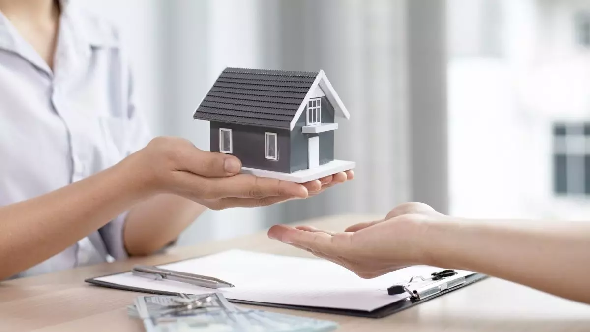 Uma pessoa entregando miniatura de casa para outra e uma prancheta com caneta sobre a mesa - Startups