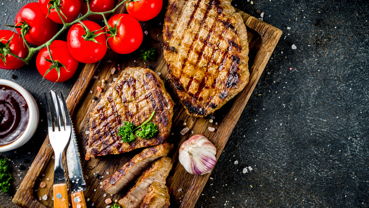 Imagem de carnes sobre uma tábua para corte, com talheres, tomates e alho - Startups