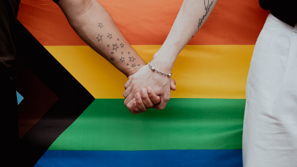 Uma mão apertando a outra e no fundo uma bandeira que simboliza a LGBTQIA+ | Startups